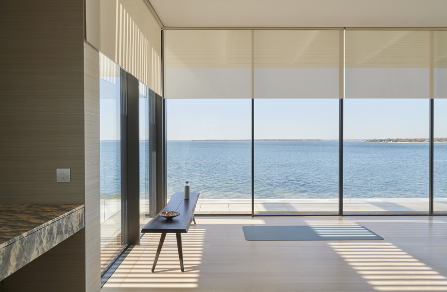 Motorized Lutron shades partially lowered over floor-to-ceiling windows in a modern waterfront home with ocean views. Social Media Bl